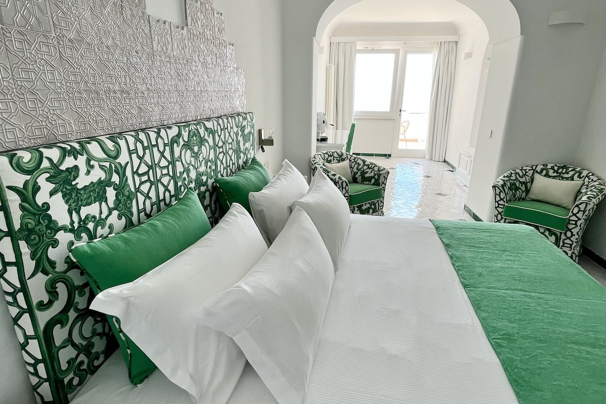 A modern hotel room with a large bed, green and white patterned headboard, and a view of the outdoor area through an arched doorway.