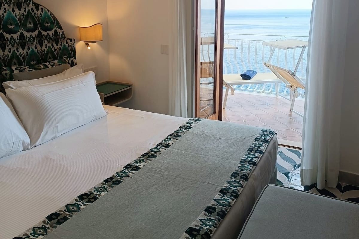 A hotel room with a bed, a nightstand, a lamp, and a view of the sea through a sliding glass door.