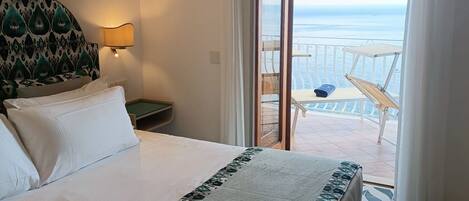 Quarto de hotel com cama, mesa de cabeceira, abajur e vista para o mar através de uma porta de vidro deslizante.