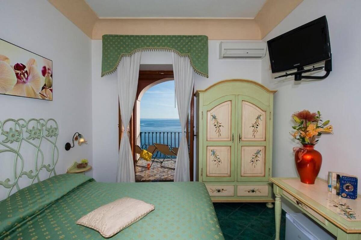 A bedroom with a bed, a desk with a vase, a television, and a view of the sea through a doorway.