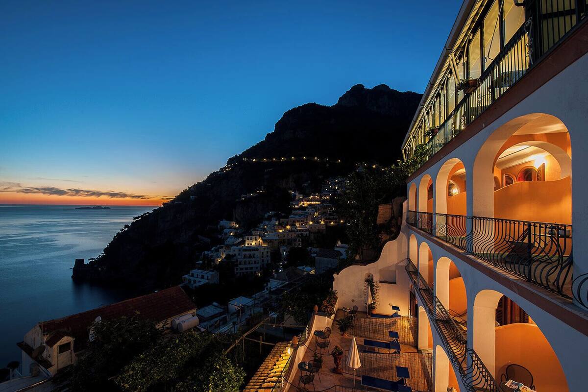 A coastal hotel with multiple balconies, a terrace with outdoor seating, and a view of the sea and a mountainous landscape.
