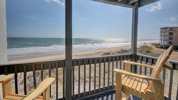 A beach view from a covered porch with wooden chairs, a building in the background, and a clear sky. - Experience oceanfront living at Shipwatch Villas 1209 in North Topsail Beach! (North Topsail Beach)