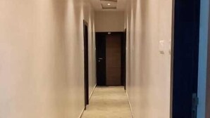 Um corredor comprido e estreito, com paredes bege e chão de azulejos, que termina numa porta escura.