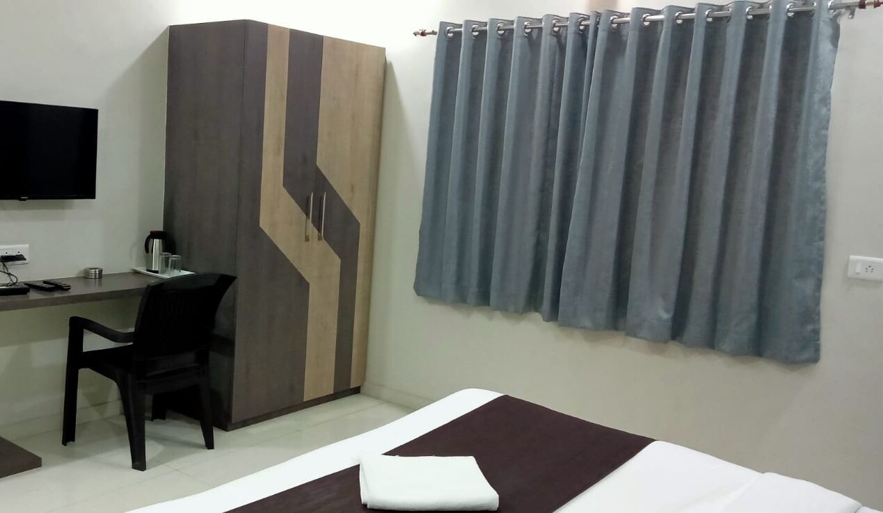 Quarto de hotel com cama, escrivaninha, cadeira, televisão e guarda-roupa.