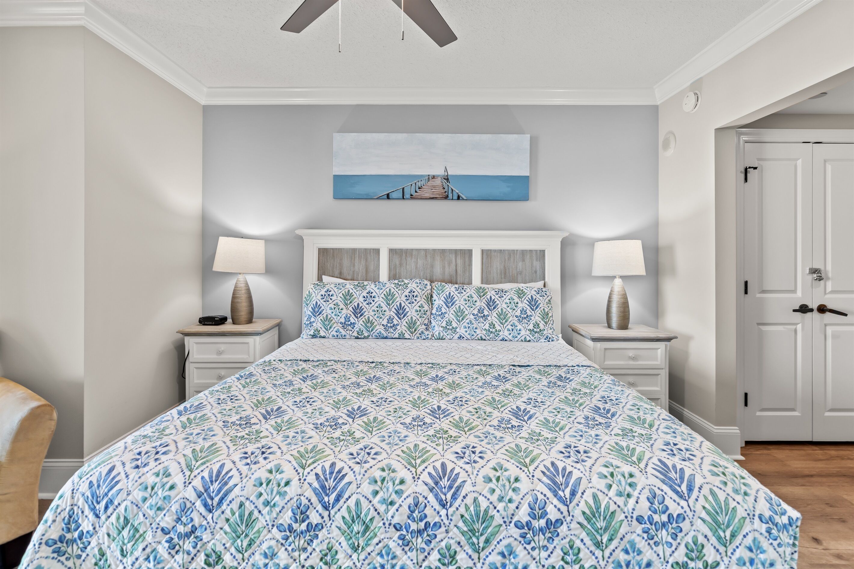 A bedroom with a large bed, two bedside tables with lamps, a ceiling fan, a painting of a pier, and a white door.
