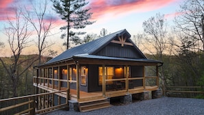 Een chalet met een houten terras en een groot raam, omgeven door bomen.