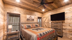 Een slaapkamer in een houten chalet, met een bed, een televisie, een plafondventilator en een houten dressoir.