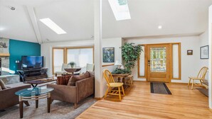 Un salon moderne avec un téléviseur, un canapé et une table basse. Il y a également un coin repas avec des chaises et une table. La pièce est dotée d’un parquet et de grandes fenêtres.