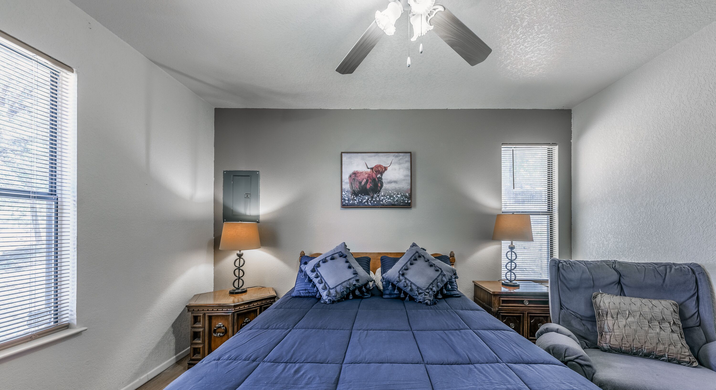A bedroom with a bed, two bedside tables, a ceiling fan, a window with blinds, and a wall-mounted light fixture.
