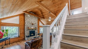 A wooden interior with a stone fireplace, a staircase, and a view of the outdoors. - Cobblestone Cottage | Dog-Friendly Escape w/ Stone Hearth & Pines (Ruidoso)