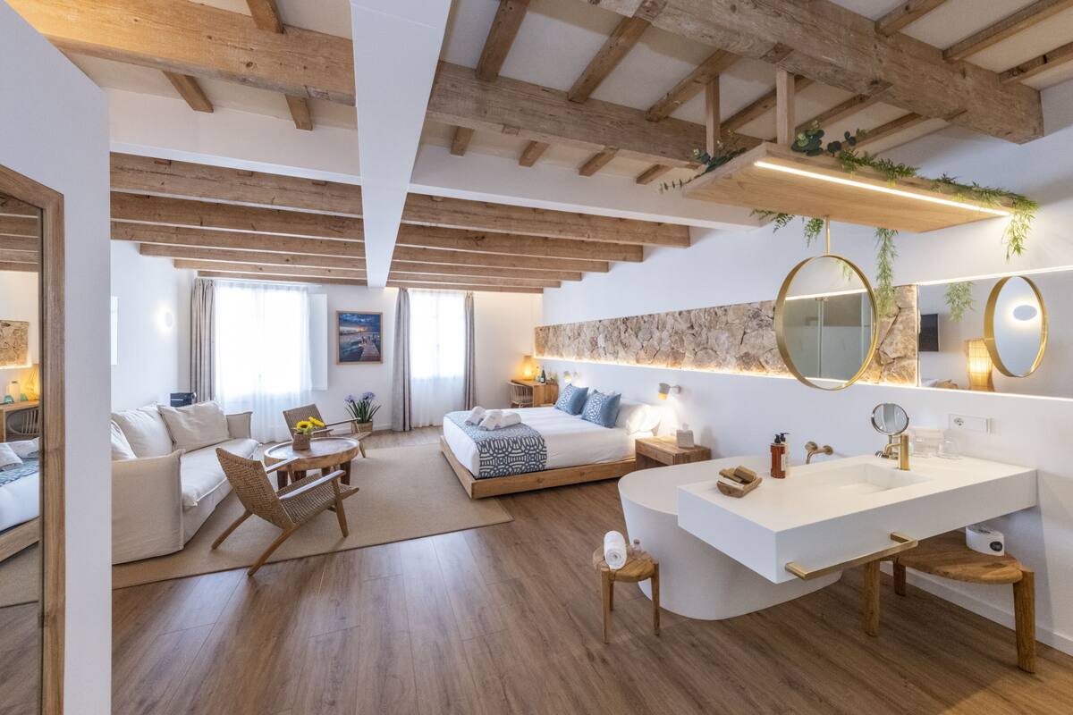 A modern hotel room with a wooden ceiling, a large bed, a white bathroom with a vanity, and a dining area with a table and chairs.