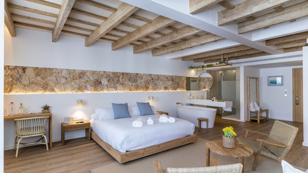 Un dormitorio moderno con pared de piedra, vigas de madera en el techo, una cama grande, mesitas de noche, un escritorio y una zona de estar con muebles de mimbre.