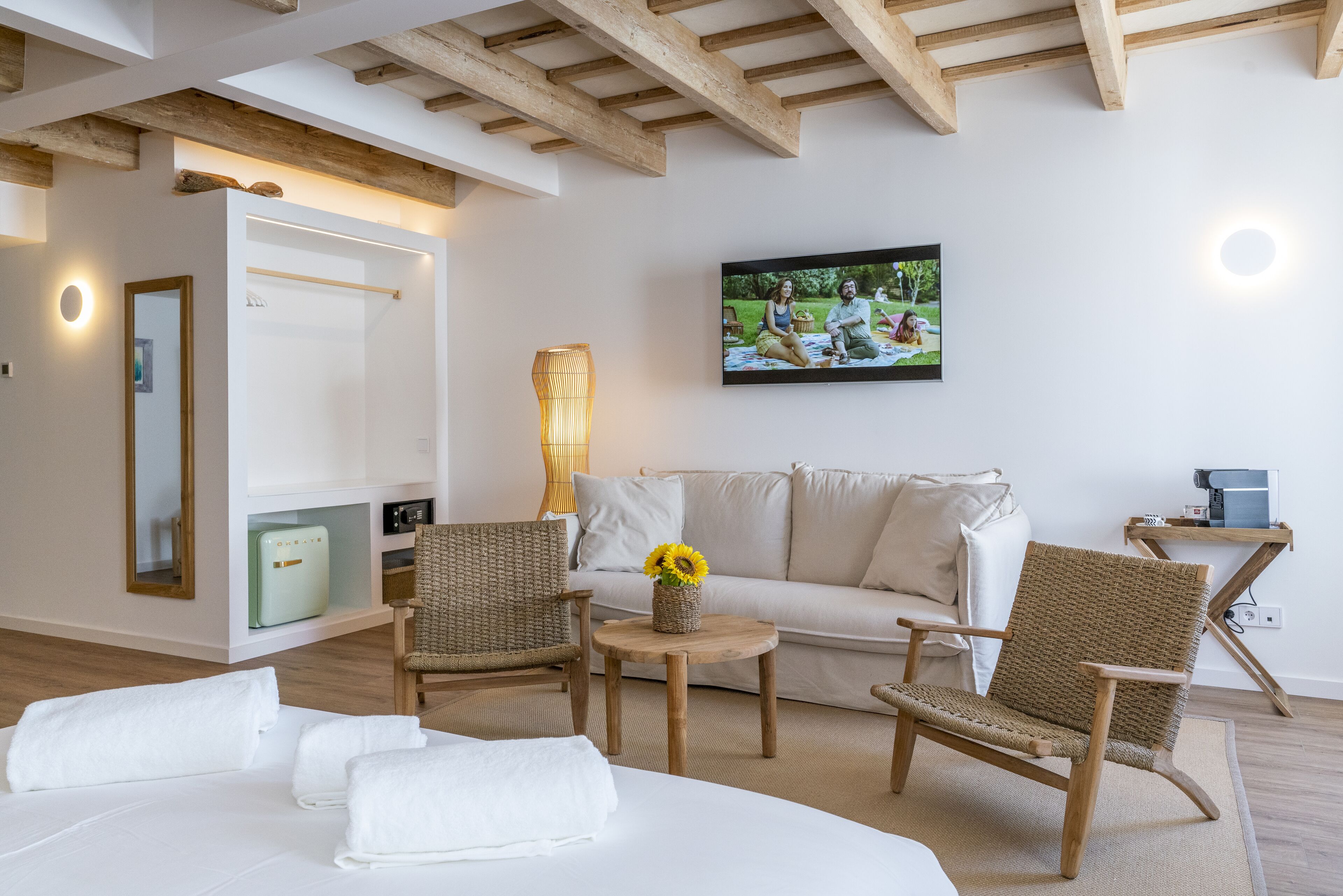 Ein modernes Hotelzimmer mit einem Bett, zwei Korbstühlen, einem kleinen Tisch mit einer Vase voller Sonnenblumen, einer Couch, einem Fernseher und einer Holzbalkendecke.