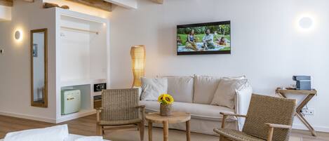 A modern hotel room with a bed, two wicker chairs, a small table with a vase of sunflowers, a sofa, a television, and a wooden ceiling.