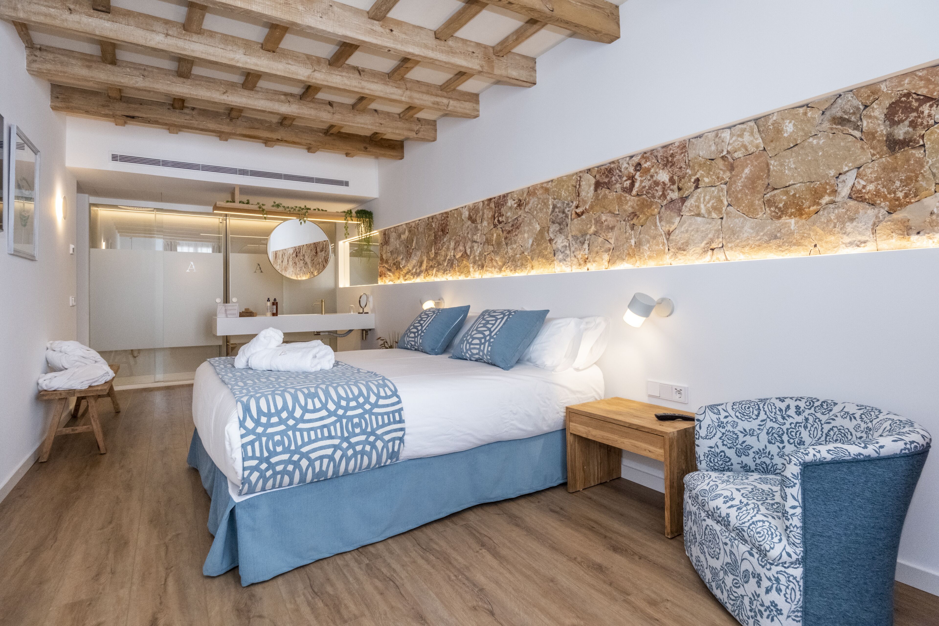 Un dormitorio moderno con una cama grande, techo de madera, pared de piedra, una mesita de noche, una silla y un espejo.