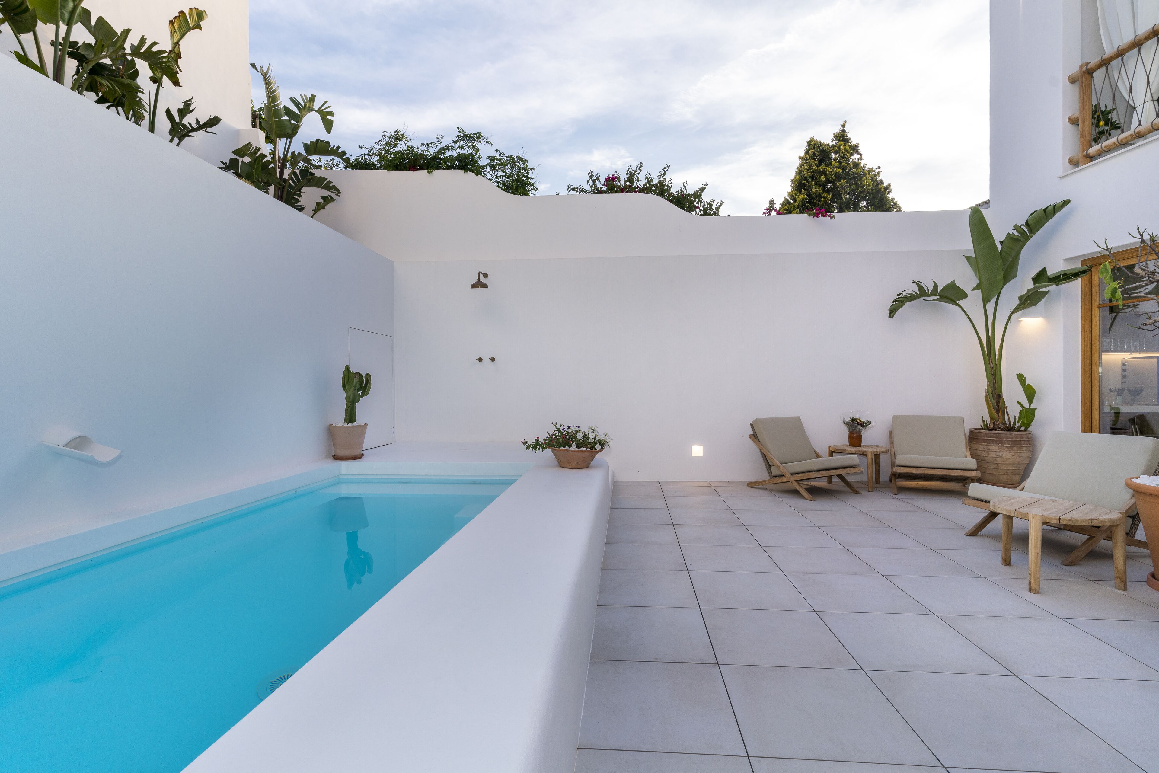 Un área de piscina moderna al aire libre con una piscina azul cristalina, rodeada de muros blancos y plantas en macetas. Hay sillas de madera para lounge y una mesita con una planta.