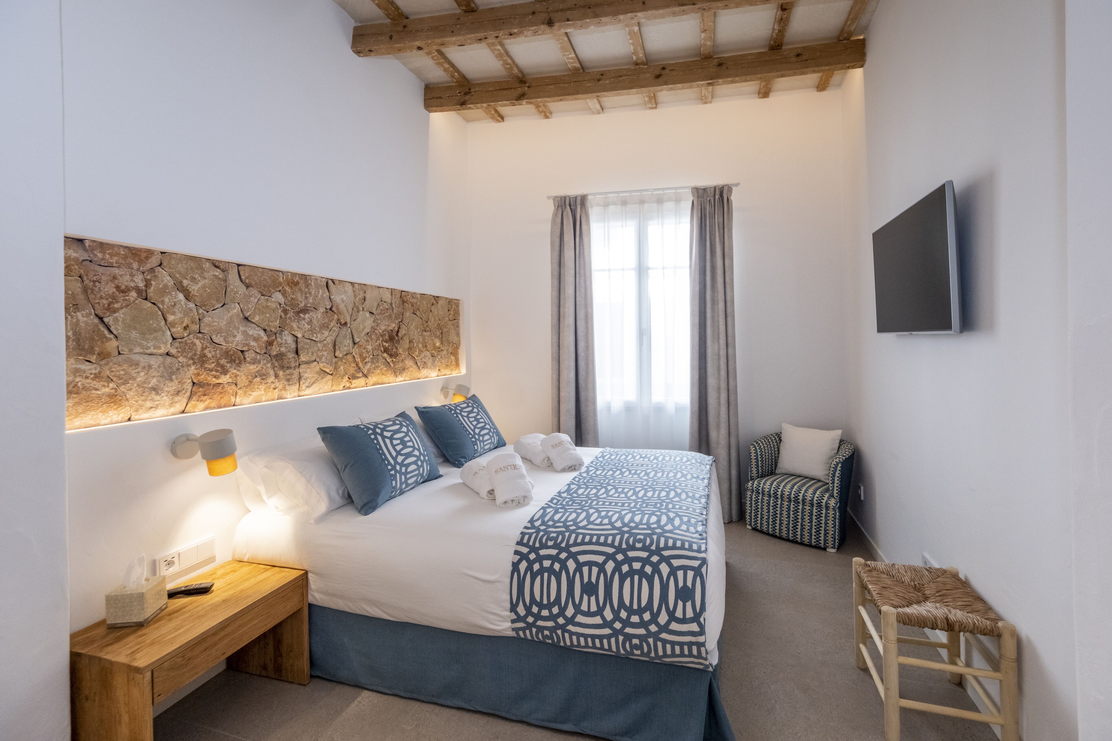 Un dormitorio con pared de piedra, cama con ropa de cama azul, mesita de noche con lámpara, una silla y televisor montado en la pared.