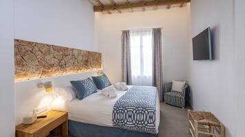 Un dormitorio con pared de piedra, cama con ropa de cama azul, mesita de noche con lámpara, una silla y televisor montado en la pared.