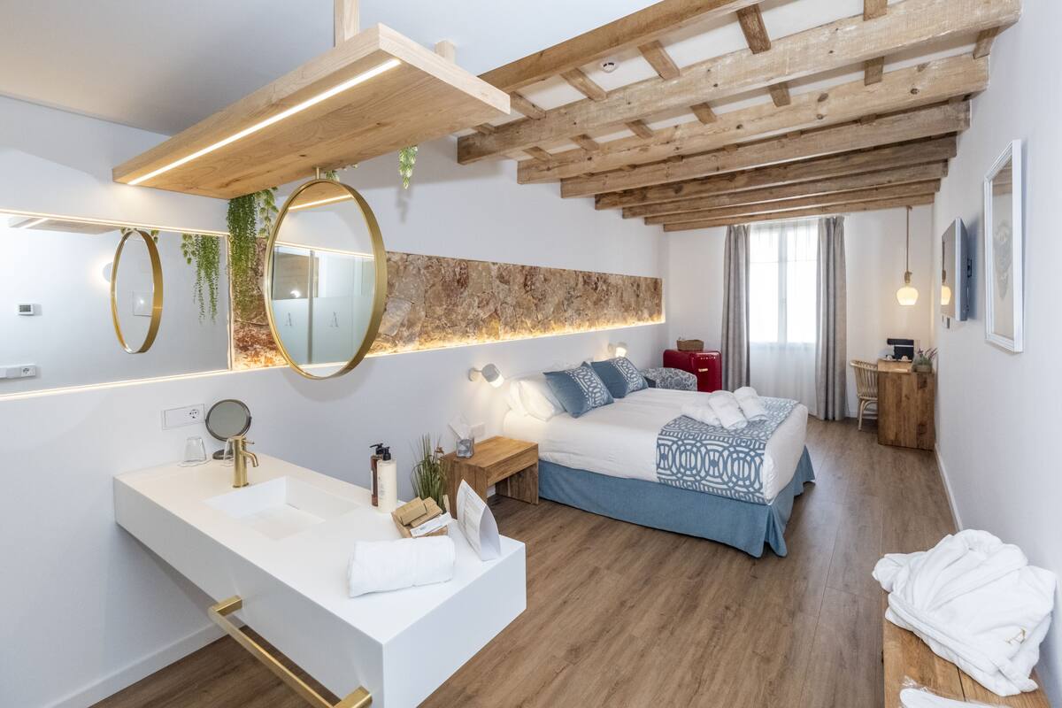 A modern bedroom with a wooden ceiling, a large bed with blue bedding, a white vanity with a gold faucet, and a large mirror.