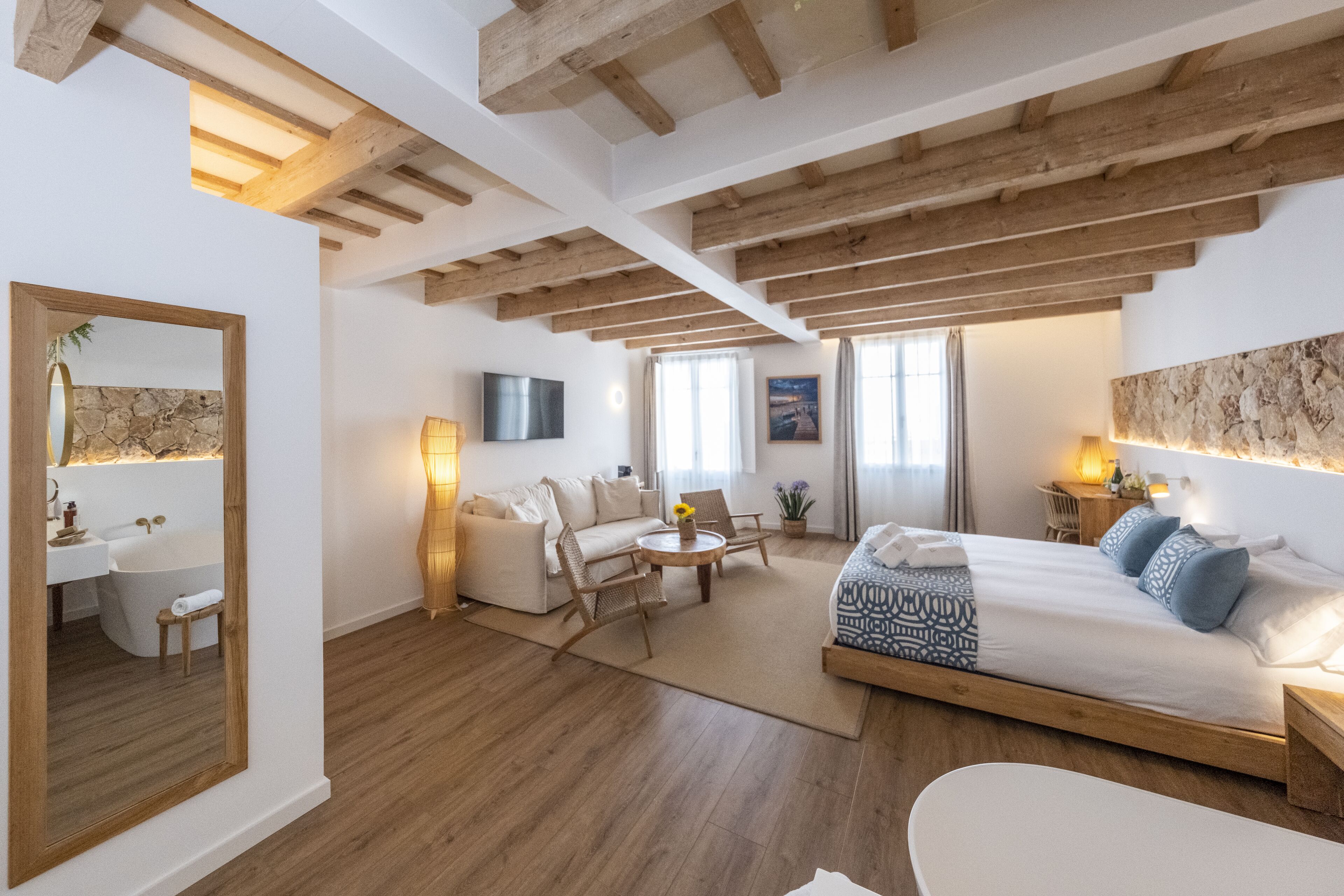 Una habitación amplia con techo de madera, una cama grande con ropa de cama estampada, un sofá, una pequeña mesa con sillas, un televisor y una pared de piedra.