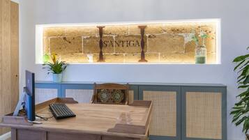 Una mesa de comedor de madera con una computadora y un teclado, una pared decorativa con las letras 'S'ANTIGA', y una ventana con plantas.