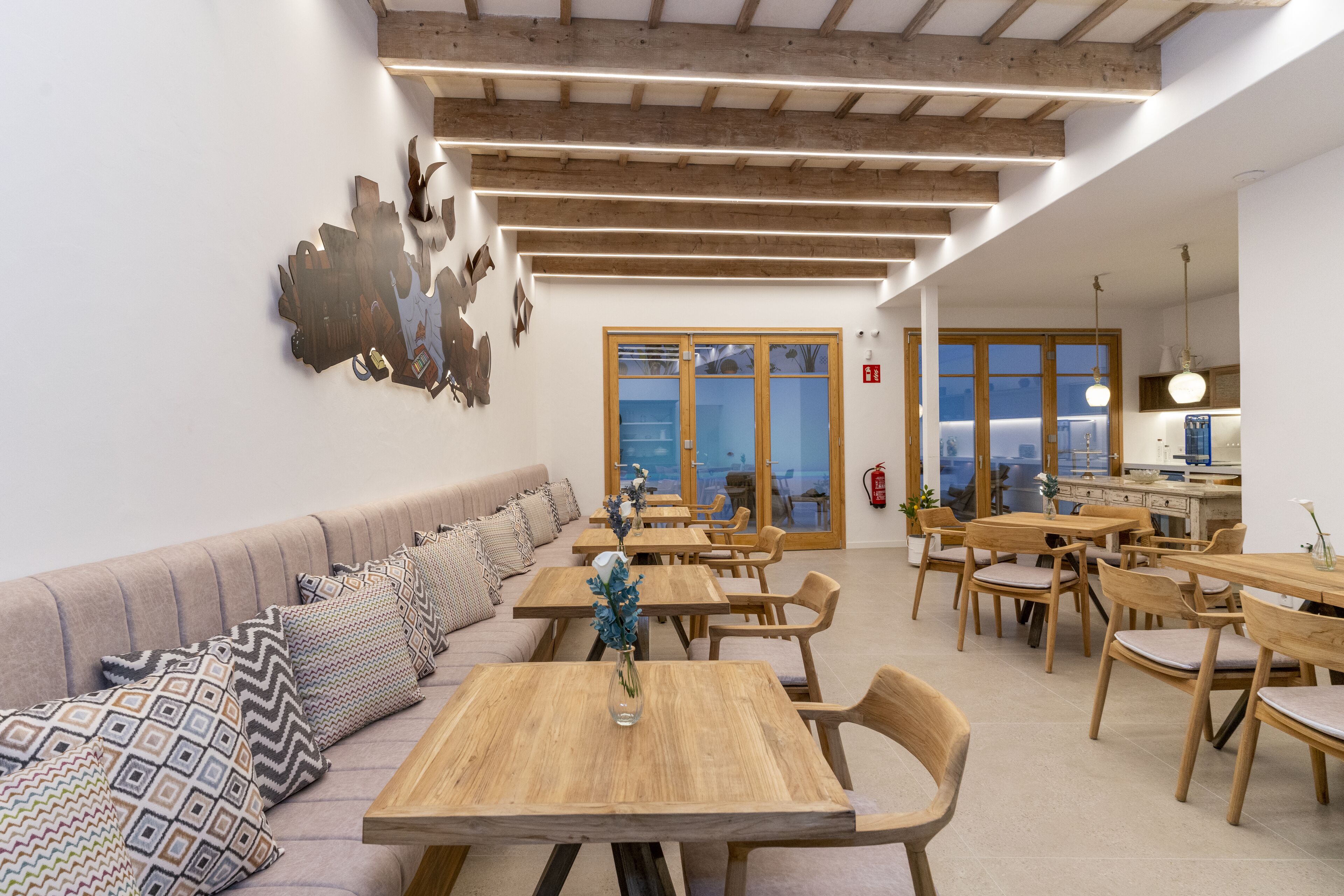 A modern dining area with wooden tables and chairs, a sofa with patterned cushions, and a large abstract wall art piece.