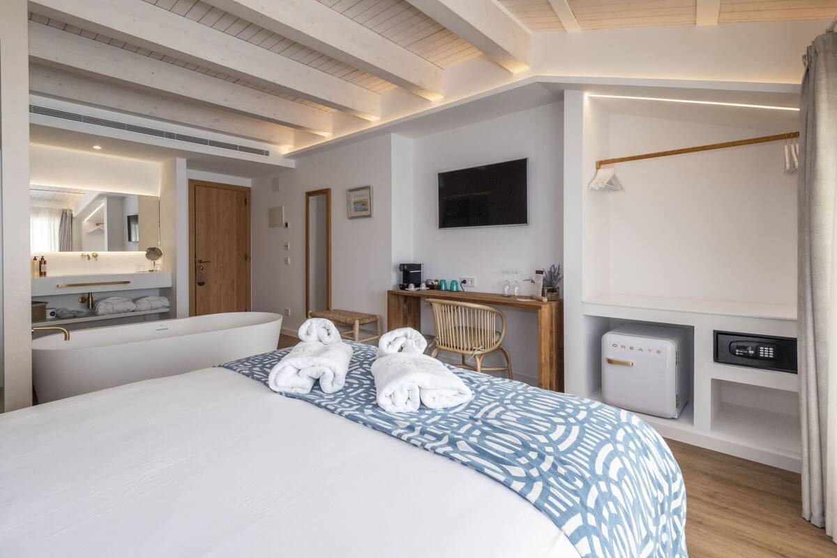 A hotel room with a large bed, a desk, a chair, a TV, a safe, and a bathroom with a mirror and towel rack.