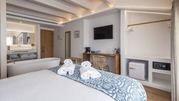 A hotel room with a large bed, a desk, a chair, a TV, a safe, and a bathroom with a mirror and towel rack.
