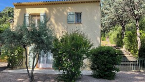 A two-story building with a beige facade, a tiled roof, and a small porch. There are two windows with shutters, and a tree and shrub in front. - Provençal villa (Le Thoronet)