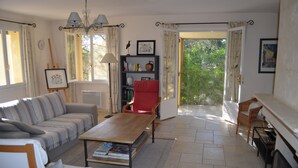 A living room with a sofa, a red chair, a coffee table with books, a bookshelf, and a view of greenery through an open door. - Provençal villa (Le Thoronet)