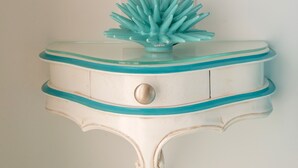 Une table d’appoint blanche, fixée au mur, à deux niveaux, avec des bordures turquoise et un seul tiroir, sur laquelle repose un objet décoratif turquoise.