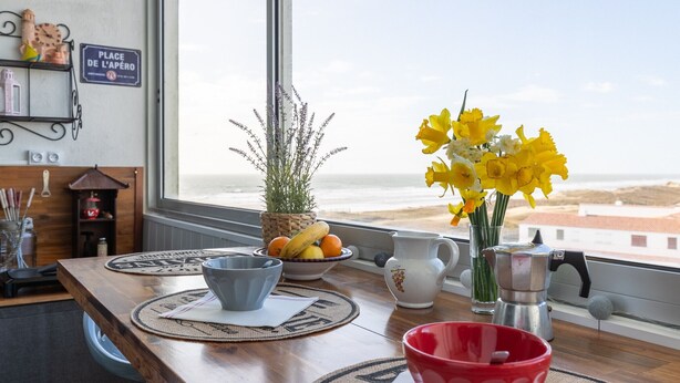 Een keuken met uitzicht op het strand, een tafel met een fruitschaal, een bloemenvaas en een koffiezetapparaat.
