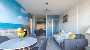 Een woonkamer met uitzicht op het strand, een eethoek met tafel en stoelen, en een keuken op de achtergrond.