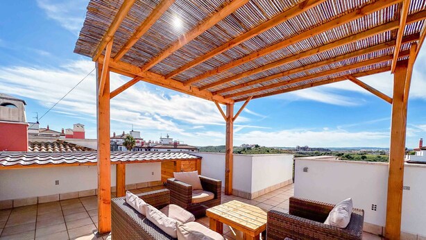 A covered outdoor seating area with wicker furniture and a wooden table, overlooking a cityscape.