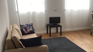Un salon avec un canapé beige, une petite table ronde, un tapis foncé et un téléviseur posé sur un meuble.