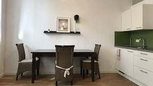 Un coin repas avec une table en bois foncé, des chaises en osier, un luminaire suspendu, un tableau encadré au mur et un pare-feu vert dans la partie cuisine.