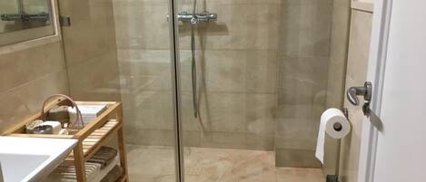 A modern bathroom with a glass-enclosed shower, a wooden shelf with towels, and a toilet with a paper towel holder.