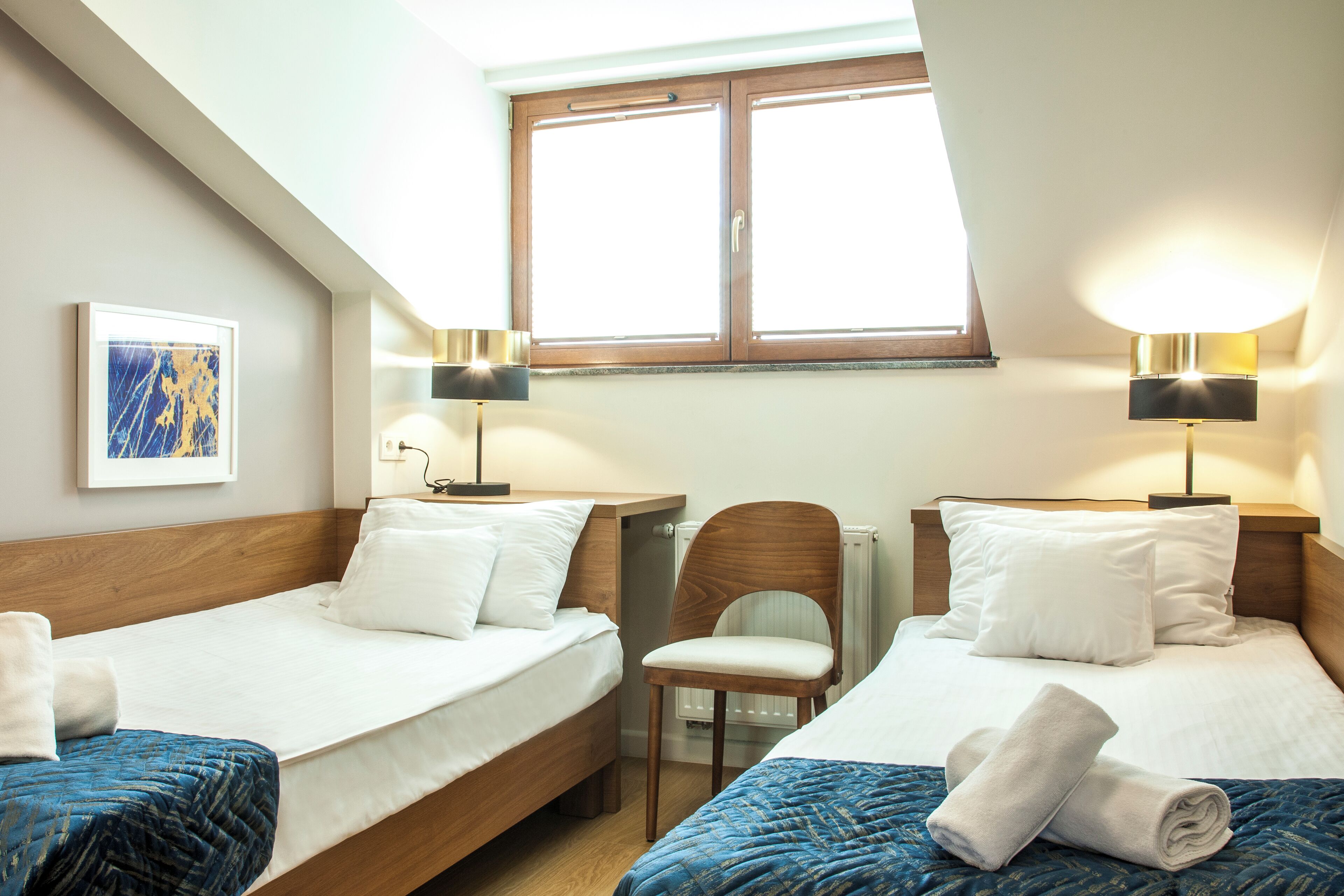 A hotel room with two beds, a chair, a window, and a painting on the wall.