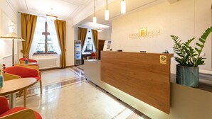 A hotel reception area with a wooden reception desk, a plant, and a window with curtains. - Portal House Apartments (Kraków)