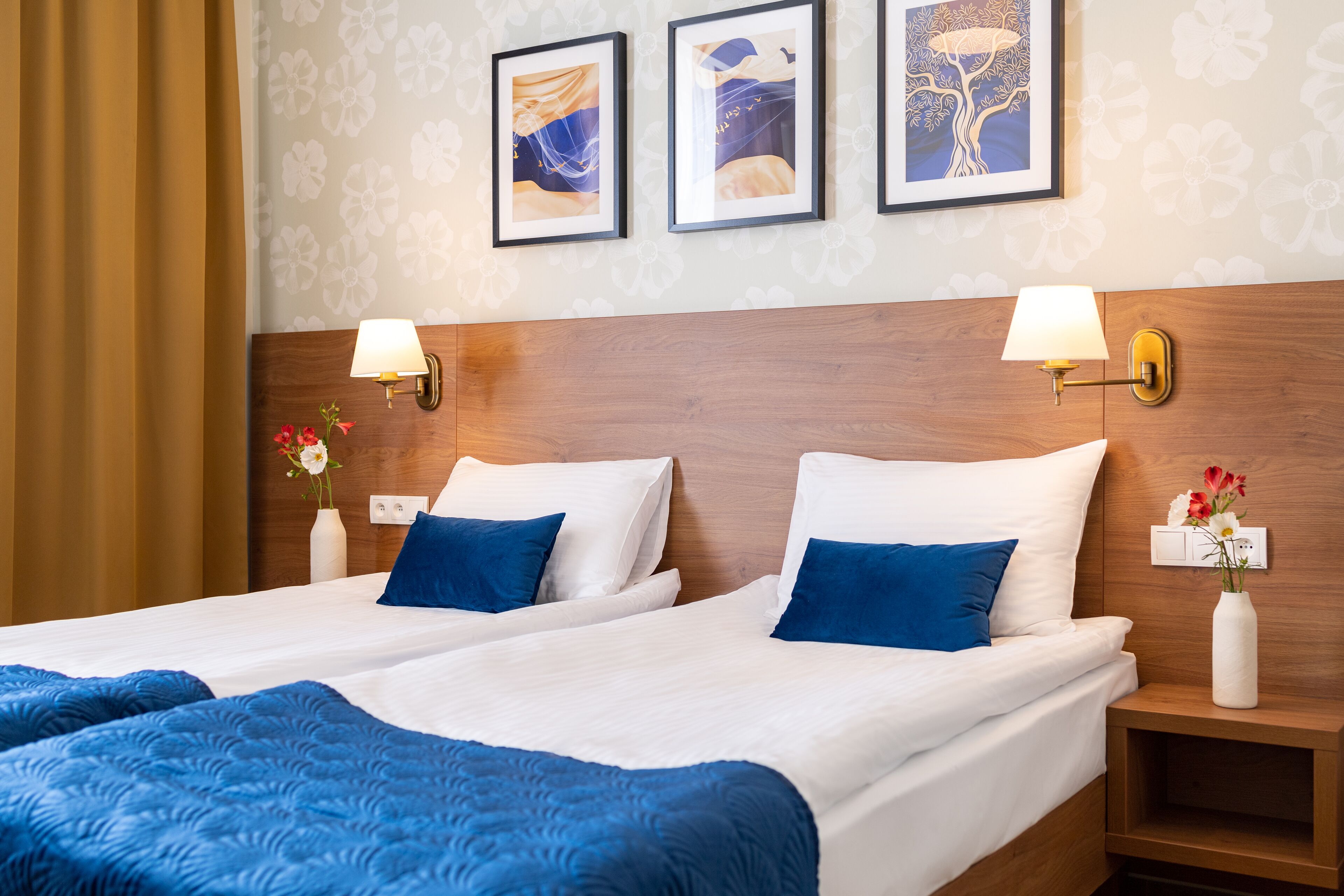 Un lit double avec une literie blanche et bleue, deux lampes fixées au mur et trois œuvres d’art encadrées accrochées au mur.