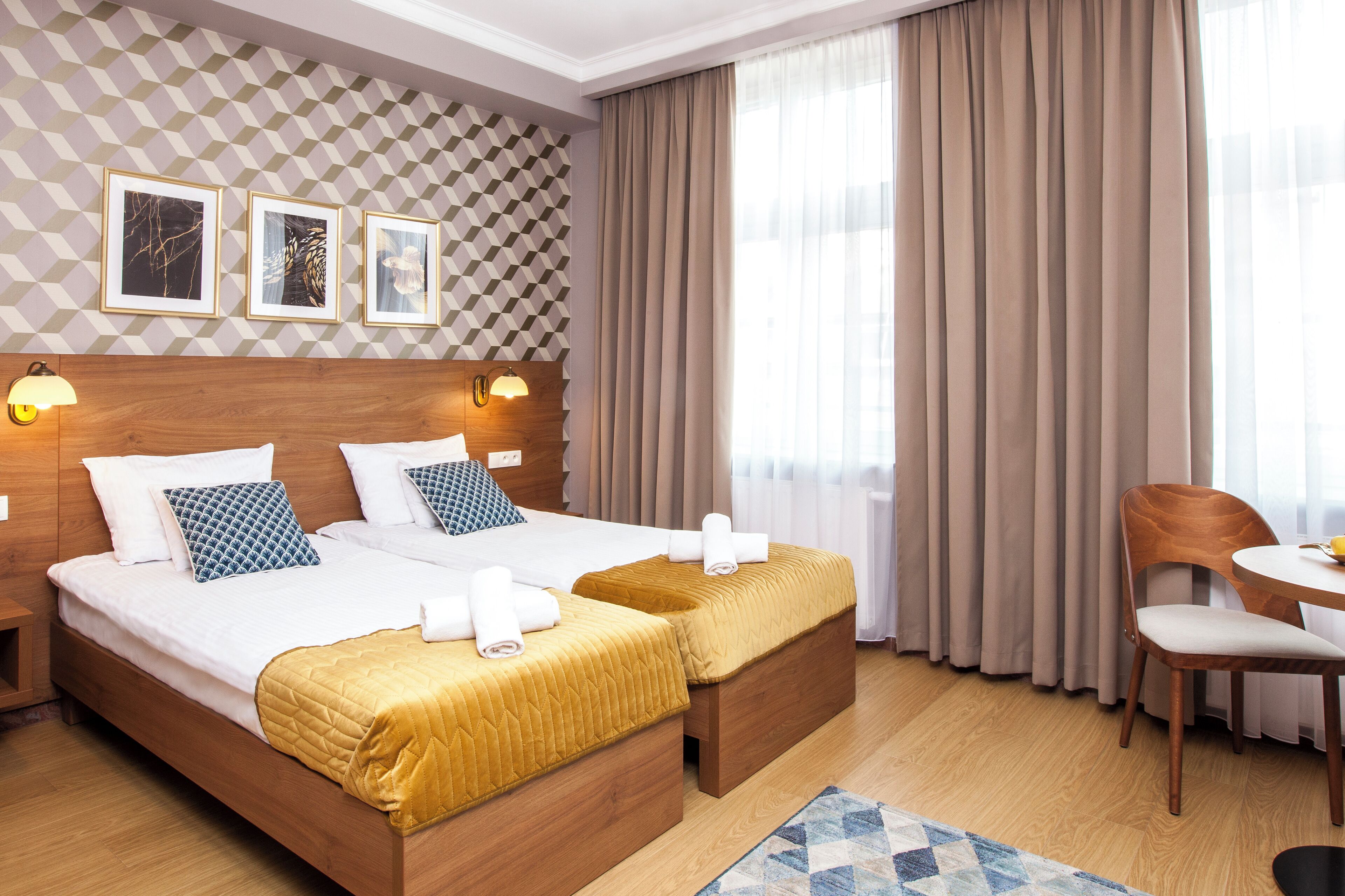 Una habitación de hotel con cama de madera, dos mesitas de noche, una mesa redonda con una silla, pared con diseño y un ventanal con cortinas.