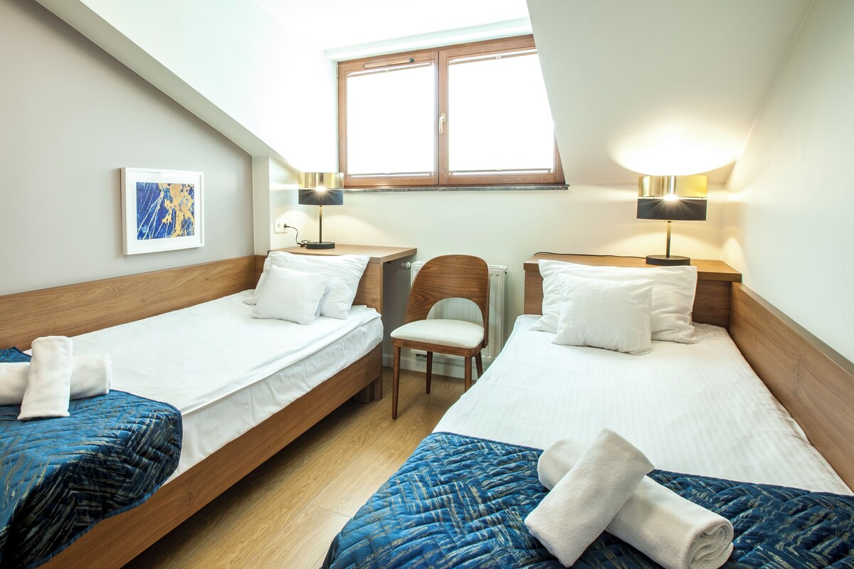 A hotel room with two single beds, a wooden headboard, a small chair, a framed picture on the wall, and a window with natural light.
