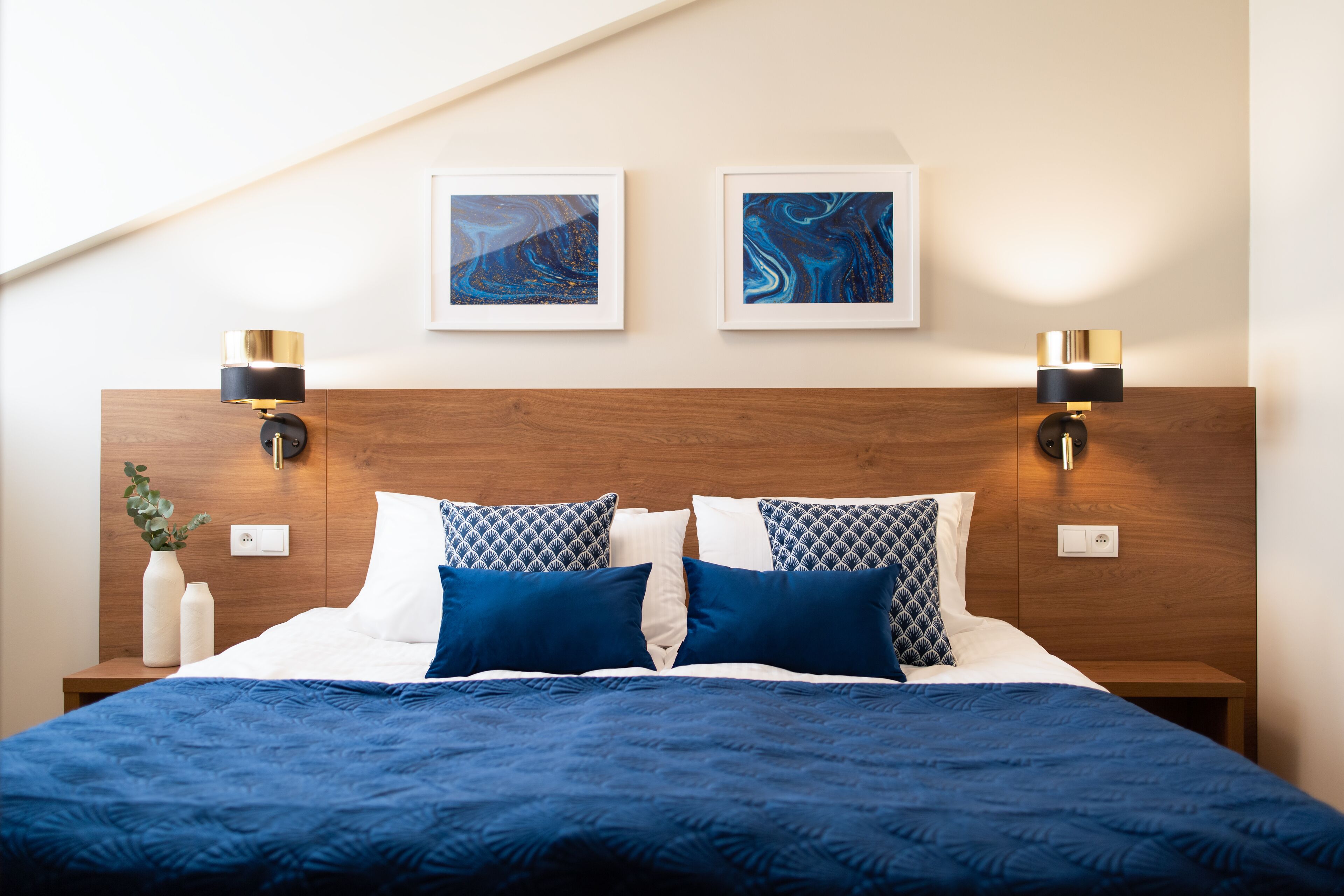 A neatly made bed with a blue quilt, two wall-mounted lamps, and two framed artworks on the wall.