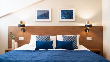 A neatly made bed with a blue quilt, two wall-mounted lamps, and two framed artworks on the wall.