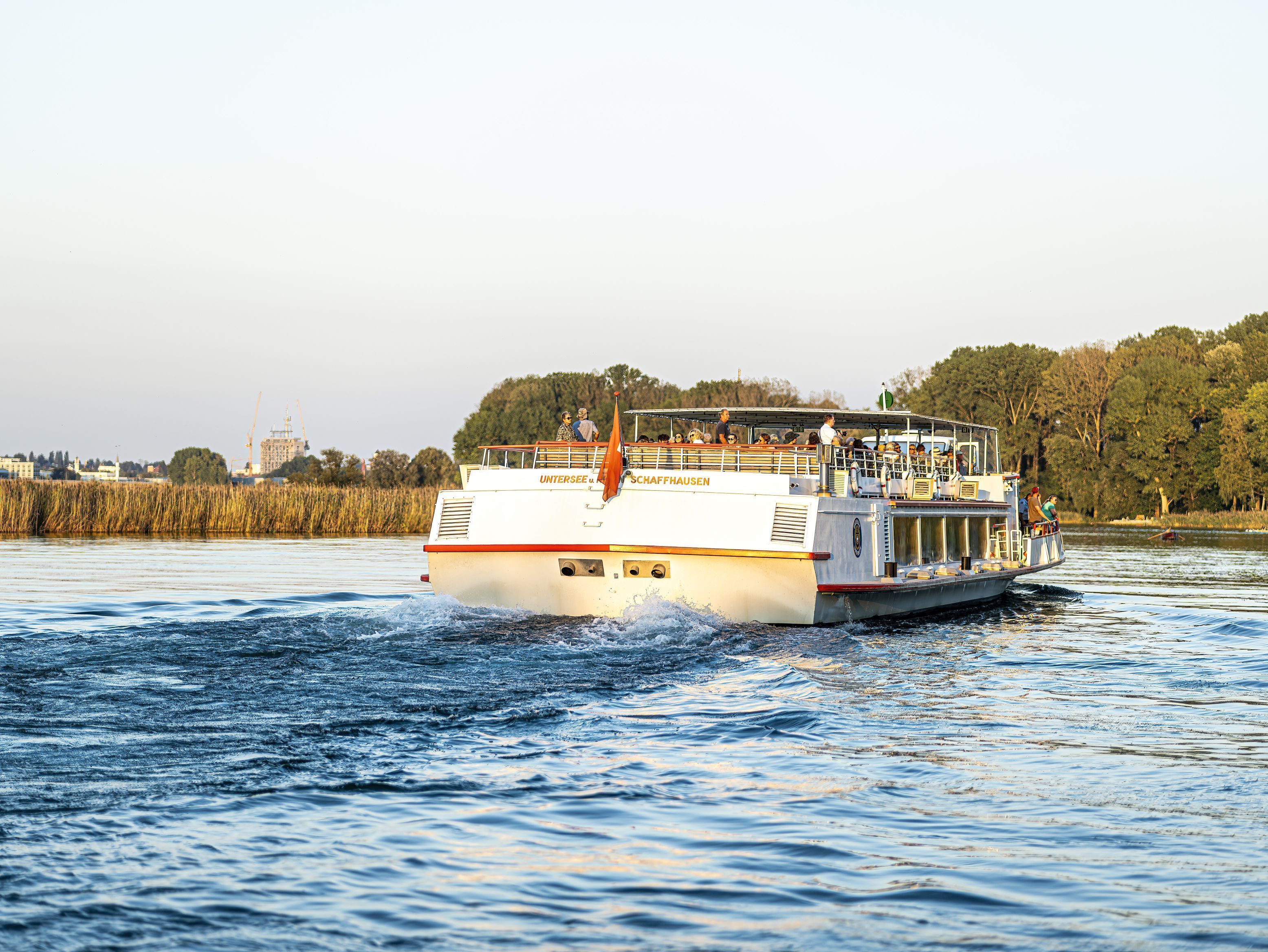 A riverboat with a flat deck and a covered upper deck, passengers seated, and a flag at the stern.