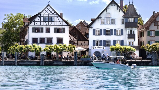 A traditional European town with historic buildings, a boat docked at the waterfront, and people enjoying the area. - Hotel Porto Sofie (Gottlieben)