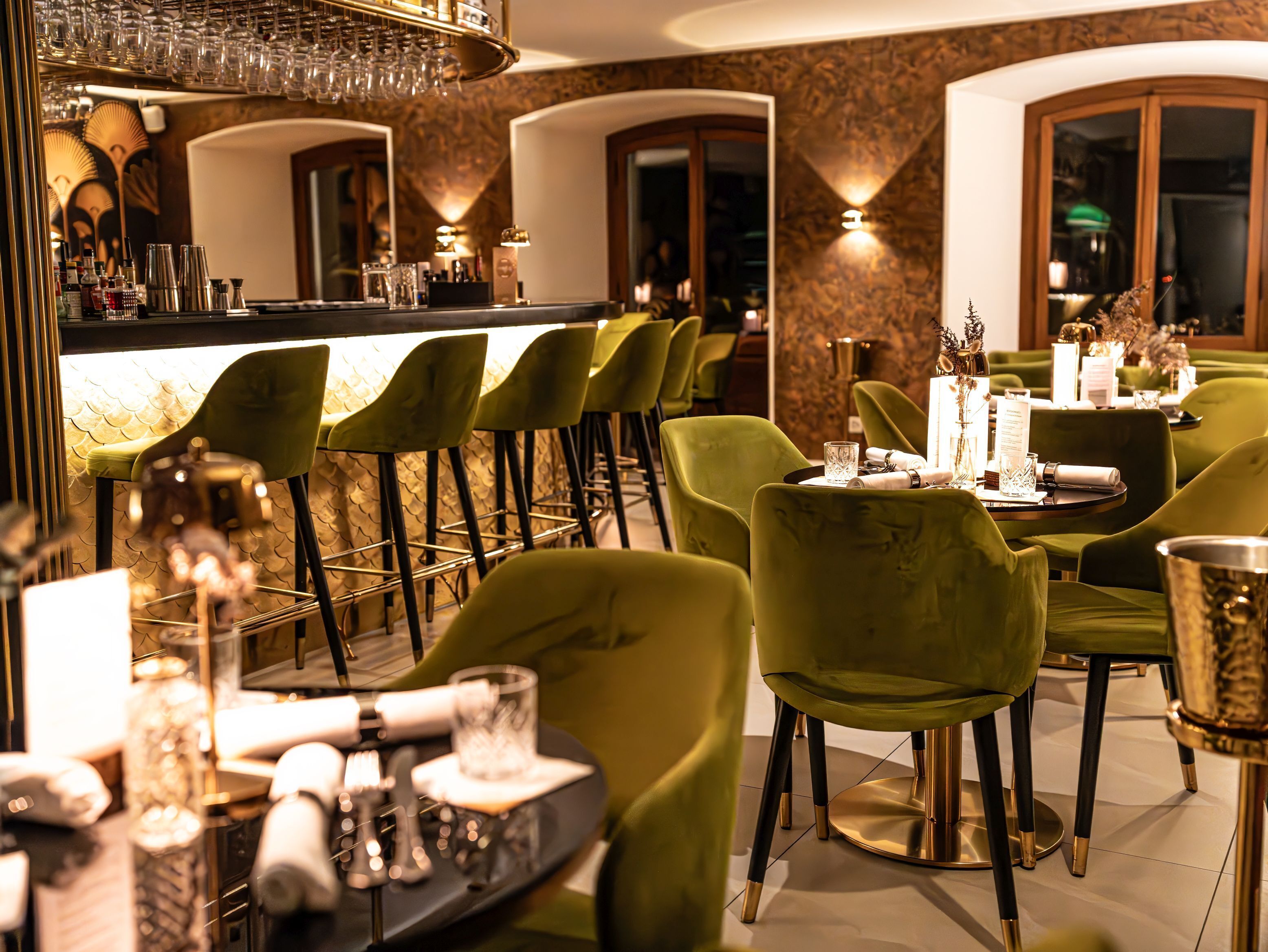 Een restaurant met groen bekleden stoelen, een bar met glazen en een goed verlicht interieur.