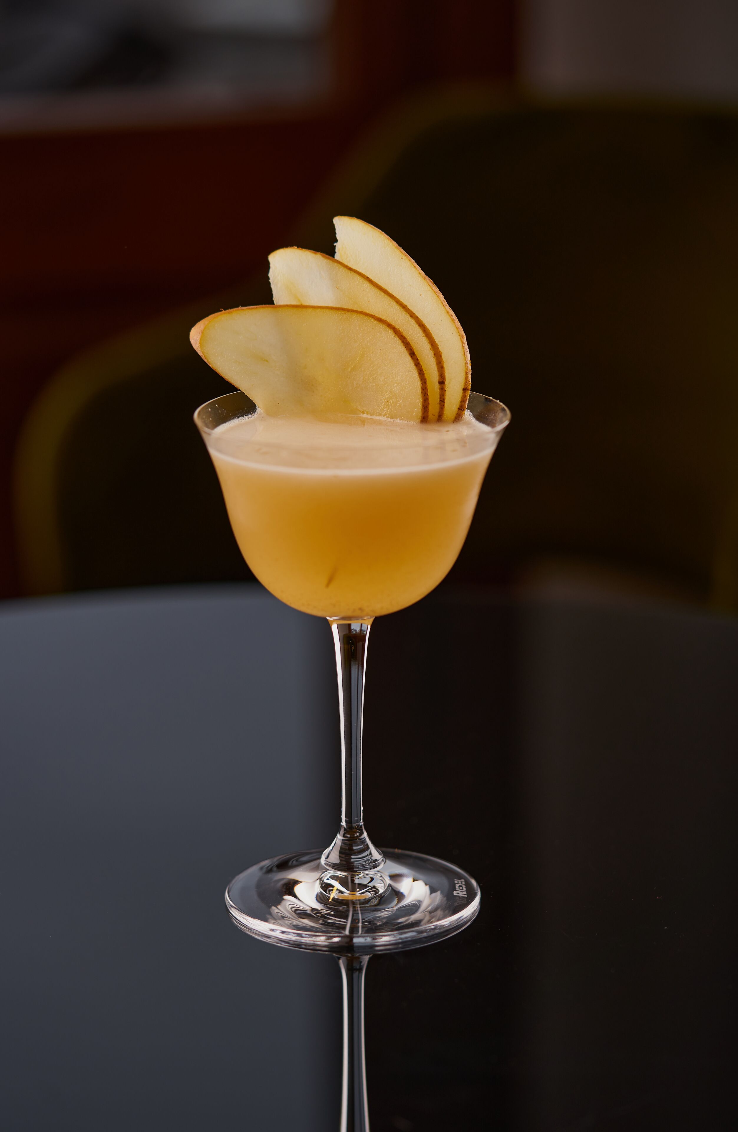 A cocktail glass with a clear stem and a wide base, filled with a light-colored beverage and garnished with apple slices.