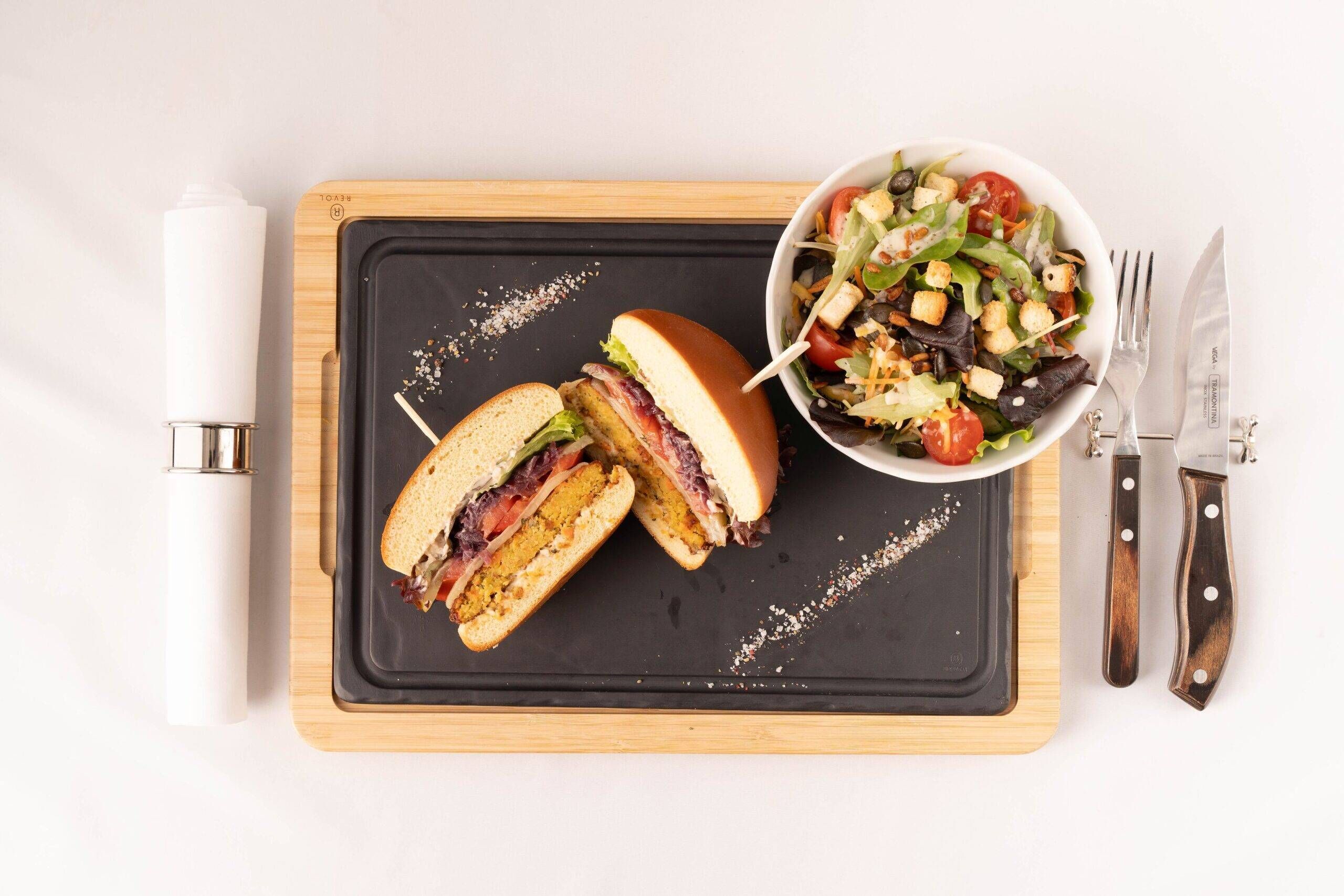 A sandwich cut in half on a black slate board, a bowl of salad, a white napkin dispenser, and a knife and fork set on a wooden board.