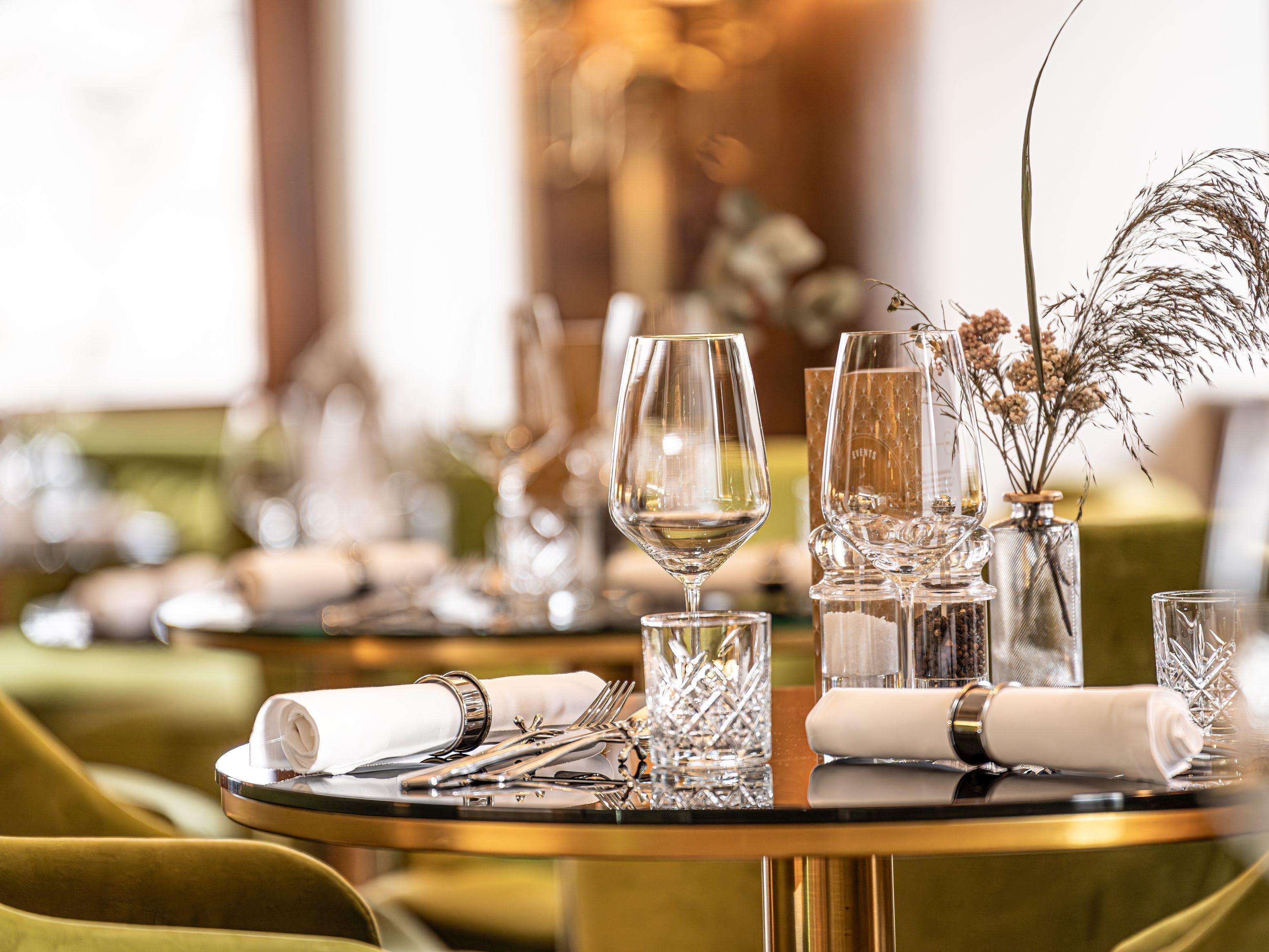Une table à manger dressée avec des verres à vin, des couverts et un vase contenant des fleurs séchées.